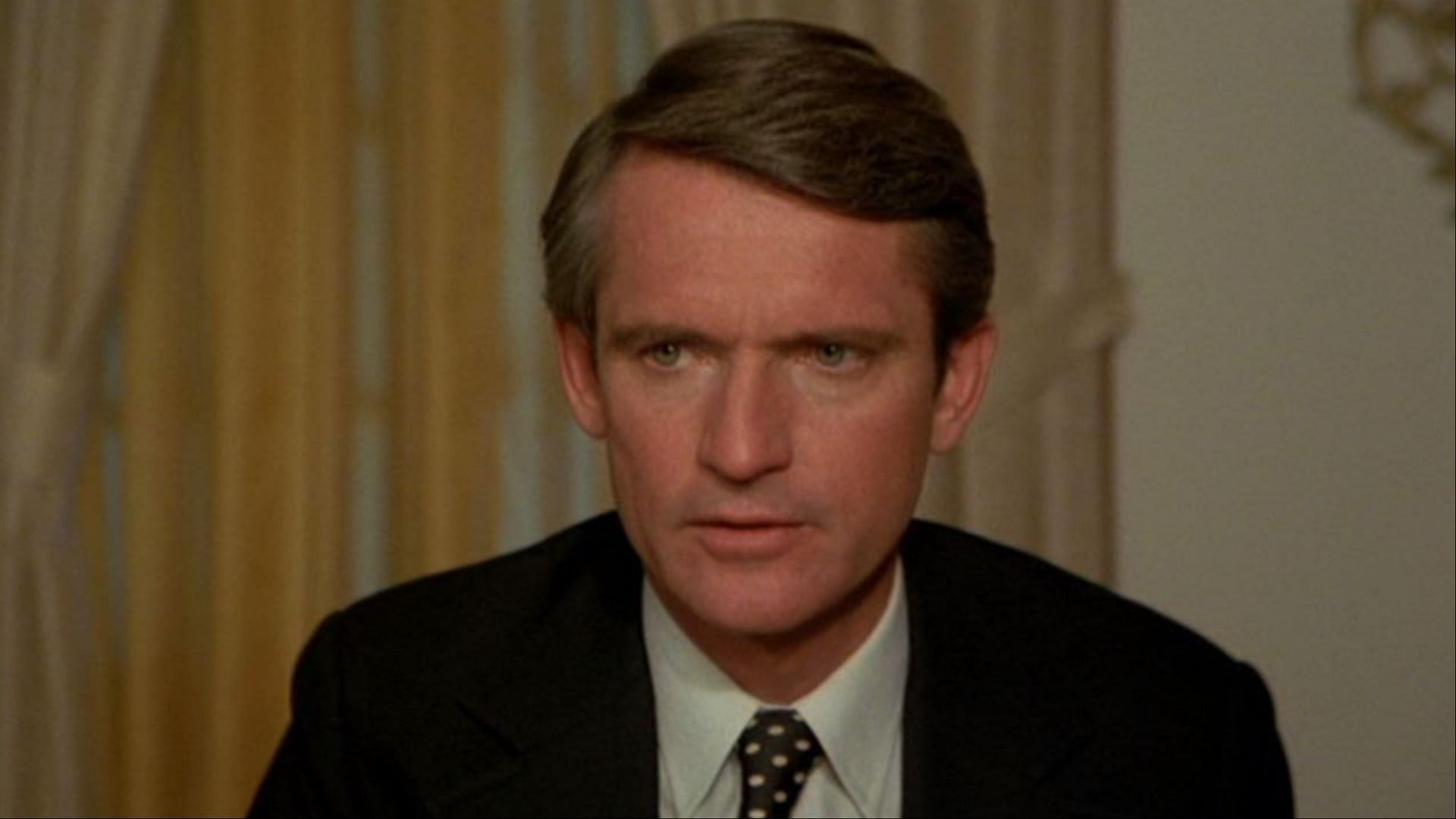Chris Robinson in a tense moment from a 1970s film, wearing a black suit and tie. (Image via FawsomeTV)